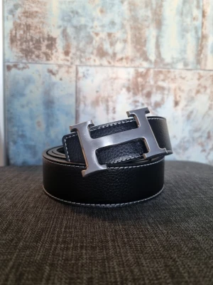 Svart skärp med H-spänne  - Snyggt svart skärp med ett stort metalliskt H-spänne framtill. Skärpet är gjort i skinn med vit kontrastsöm längs kanterna och har en stilren, klassisk design som passar till många outfits.