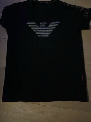 Svart t-shirt från Armani med logga - Svart t-shirt från Armani med den klassiska örnloggan i silver och grått på bröstet. T-shirten har rund halsringning och korta ärmar. Perfekt för dig som gillar stilrena märkesplagg.