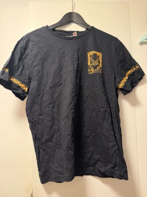 Svart Black Panther t-shirt H&M - Cool svart t-shirt från H&M med Marvel Black Panther-tryck i gult på bröstet och detaljer på ärmarna. T-shirten har rund hals och är kortärmad. Tillverkad i mjuk bomull, perfekt för dig som gillar superhjältar och streetstyle.