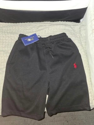 Svarta shorts från Ralph Lauren - Svarta shorts från Ralph Lauren med klassisk röd logga broderad på benet. De har elastisk midja med snörning för skön passform och är tillverkade i mjuk bomull. Perfekta för chill dagar eller när du vill ha en clean look.