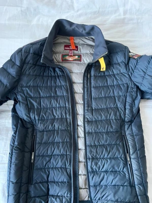 Mörkblå dunjacka från Parajumpers - Snygg mörkblå dunjacka från Parajumpers med quiltad design, ståkrage och två dragkedjeförsedda fickor framtill. Jackan har en gul detalj vid halsen och ett tydligt Parajumpers-märke på ärmen. Perfekt för kalla vinterdagar. Storlek M