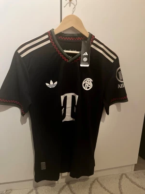 Svart Bayern München Adidas tröja - Snygg svart Bayern München matchtröja från Adidas med vita detaljer och röda inslag vid hals och ärmslut. Tröjan har klassiska Adidas-ränder på axlarna, klubbmärke och sponsorlogga framtill samt patch på ärmen. Perfekt för fotbollsfans!
