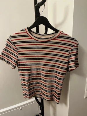 Randig ribbad t-shirt i rött och svart - Säljer en ribbad t-shirt med korta ärmar och rund hals. T-shirten har ett randigt mönster i vitt, svart och rött. Passformen är något tight och materialet känns mjukt och stretchigt, perfekt för en avslappnad look.