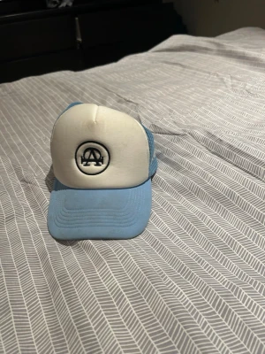 Blå och vit truckerkeps med logga - Snygg truckerkeps i ljusblått och vitt med svart broderad logga framtill. Kepsen har mesh på sidorna och bak för extra ventilation och en böjd skärm i blått. Perfekt för en avslappnad streetwear-look.  ALL CAP ONE 