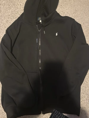 Svart hoodie från Polo Ralph Lauren - Klassisk svart hoodie med dragkedja från Polo Ralph Lauren. Har huva med dragsko, fickor framtill och den ikoniska vita logobrodyr på bröstet. Perfekt för en chill och stilren look.
