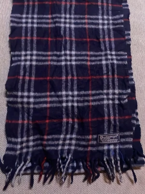 Rutig halsduk från Burberrys - Klassisk halsduk i mörkblått med rött och vitt rutmönster från Burberrys. Tillverkad i 100% lammull och har fransar i kanterna. 