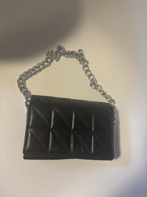 Svart väska med kedja - Snygg svart handväska i quiltat mönster med en rejäl silverfärgad kedja som axelrem. Väskan har en stilren, rektangulär form och är tillverkad i ett glansigt material som liknar skinn. Perfekt accessoar för att lyfta din outfit.
