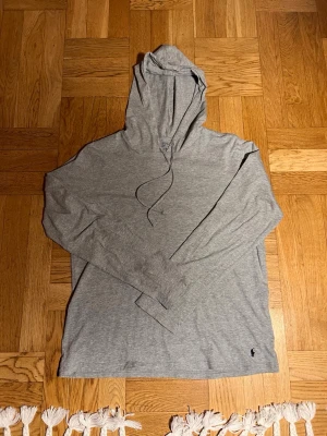 Grå hoodie från Polo Ralph Lauren - Säljer en stilren grå hoodie från Polo Ralph Lauren med klassisk huva och dragsko. Tillverkad i mjukt och tunt bomullsmaterial, perfekt för en avslappnad look. Liten broderad logga i svart vid nederkanten. Långärmad och enkel design.