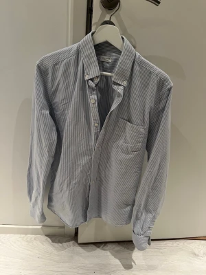 Randig skjorta från A Day's March - Snygg skjorta med smala blå och vita ränder från A Day's March. Klassisk button-down krage, bröstficka och vita knappar. Skjortan har en custom fit och är tillverkad i ett mjukt bomullstyg. Perfekt för en stilren och avslappnad look.