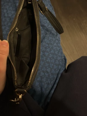 Svart axelväska i skinnimitation - Snygg svart axelväska med justerbart axelband och dragkedja upptill. Väskan har en enkel, stilren design med metall-detaljer och en liten metallbricka framtill. Perfekt storlek för det viktigaste och passar till de flesta outfits.