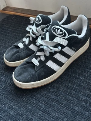 Adidas Campus Svarta  - Klassiska Adidas Campus sneakers i svart mocka med vita detaljer och de ikoniska tre ränderna på sidan. Skorna har vit sula, vita skosnören och Adidas-logga på plösen och hälen. Låg modell som passar till många olika stilar. Den grejen vid snörena har gått av, men ingen som påverkar hur den sitter eller något som syns!
