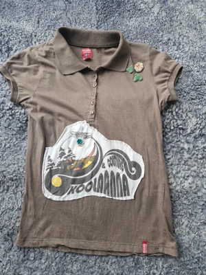 Brun Koola Anna t-shirt med tryck - Snygg brun t-shirt från Koola Anna med krage, knappar och korta ärmar. Framsidan har ett stort grafiskt tryck i svart, vitt och gult samt dekorativa virkade blommor. Fint skick förutom storlekslappen som släppt lite i sömmen