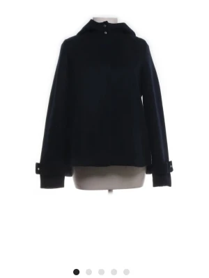 Svart hoodiejacka med knappar - Stilren svart hoodiejacka med knappar vid halsen och justerbara spännen vid ärmsluten. Jackan har en luva och är tillverkad i ett tjockare tyg som passar perfekt för kyligare dagar. Insidan av luvan har ett rutigt foder som ger en snygg detalj.