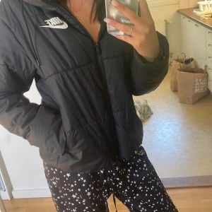 Svart pufferjacka från Nike - Svart pufferjacka från Nike med dragkedja och huva. Jackan har en sportig look med logga på bröstet och två fickor framtill. Perfekt för kyligare dagar när du vill ha en stilren och bekväm jacka.