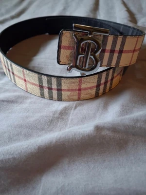 Rutmönstrat bälte från Burberry - Snyggt bälte från Burberry med klassiskt rutmönster, bältet har en svart sida och en beige sida. Bältet har ett stort metallspänne med Burberrys ikoniska monogram. Perfekt accessoar för att lyfta din outfit. 