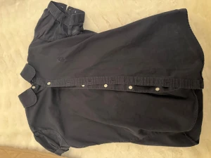 Svart kortärmad skjorta med knappar - Kortärmad svart skjorta med klassisk krage och knäppning framtill. Skjortan har en diskret broderad logga på bröstet och är tillverkad i ett mjukt bomullstyg. Perfekt för en avslappnad och stilren look.