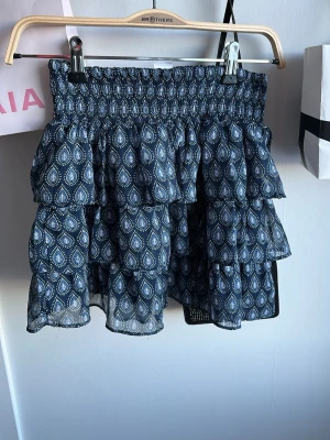 Blå volangkjol med mönster - Supersöt blå kortkjol med volanger och elastisk midja. Kjolen har ett vitt mönster och är gjord i ett lätt, luftigt material som passar perfekt till varmare dagar. Snygg att styla med en enkel topp eller stickad tröja.