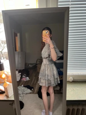 Kort omlottklänning med volang - Snygg kort omlottklänning med volangdetaljer och v-ringning. Klänningen har ett svartvitt mönster, korta ärmar och markerad midja. Perfekt för dig som gillar en feminin och trendig stil.