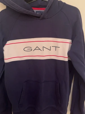 Marinblå hoodie från GANT - Snygg marinblå hoodie från GANT med vit panel och röda linjer över bröstet samt stor GANT-logga. Klassisk känguruficka framtill och huva. Perfekt för en avslappnad och trendig stil. Mjuk bomullsblandning som känns skön mot huden.