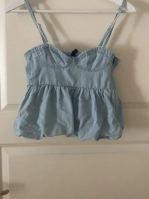 Ljusblå jeanstopp med volang - Supersöt ljusblå topp med volang och smala axelband. Toppen har en markerad byst och en luftig, flowy passform som ger en romantisk vibe. Säljer då den var för liten i storlek. Öppen för prisförslag då jag vill sälja av så fort som möjligt!💞