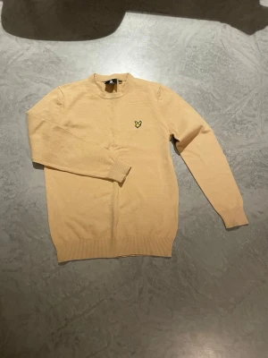 Beige stickad tröja från Lyle & Scott - Säljer en beige stickad tröja från Lyle & Scott med deras klassiska gula örnlogga broderad på bröstet. Tröjan har rund halsringning, långa ärmar och ribbade muddar vid ärmslut och nederkant. Perfekt för dig som gillar stilren och enkel design.