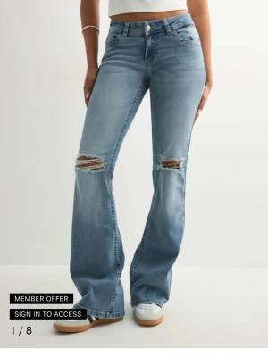 Lowwaist nelly jeans - Säljer ett par ljusblå bootcut jeans med slitningar över knäna och detaljer på bakfickorna. Jeansen har låg midja och femficksdesign. Perfekta för en avslappnad och trendig look. Endast använda en gång, nypris 699