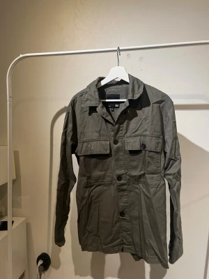 Olivgrön overshirt från 157 - Stilren olivgrön overshirt från 157 med klassisk krage och två stora bröstfickor med knapp. Skjortan har raka ärmar och knäpps framtill med svarta knappar. Perfekt lager-på-lager-plagg i slitstarkt bomullstyg.