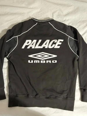 Svart stickad tröja Palace x Umbro - Svart stickad tröja från Palace x Umbro med vit logga och text på ryggen. Tröjan har vita kontrastsömmar och ribbade muddar vid ärmslut och nederkant. Perfekt för dig som gillar streetwear och sportiga vibes.