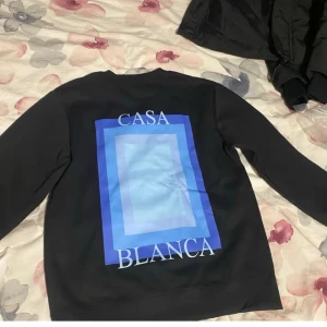  sweatshirt från Casablanca - Svart sweatshirt från Casablanca med blått grafiskt tryck och texten 'Casa Blanca' på både fram- och baksidan. Tröjan har rund halsringning och långa ärmar. Perfekt för en trendig och avslappnad stil.