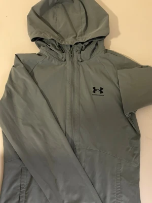 Grå vindjacka från Under Armour - Snygg grå vindjacka från Under Armour med huva och hel dragkedja framtill. Jackan har en minimalistisk design med svart logga på bröstet och praktiska sidfickor. Perfekt för blåsiga dagar och sportiga outfits.
