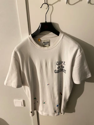 Vit Gallery Dept t-shirt med tryck - Vit t-shirt från Gallery Dept. med svart texttryck 'DÉPT. de la GALERIE' på både bröstet och ryggen. Unik design med färgstänk i olika färger. Klassisk rund hals och korta ärmar. Perfekt för dig som gillar streetwear och statement-plagg.