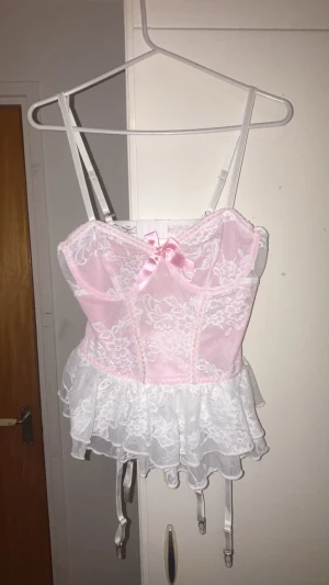 Ljusrosa & Vit Korsett - Supersöt korsett (lös) i ljusrosa och vitt med spetsdetaljer, volangkant och justerbara axelband. Framtill sitter en liten rosett i satin och hela korsetten har blommigt spetsmönster. Perfekt för dig som gillar romantisk och feminin stil. M men passar som S, kuporna vet jag riktigt inte.