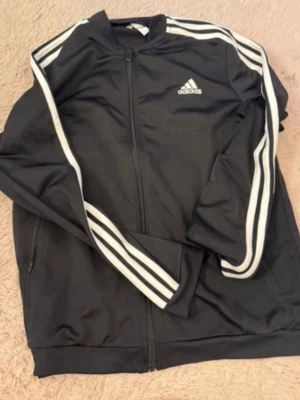 Svart Adidas ziptröja med ränder - Säljer en svart ziptröja från Adidas med klassiska vita ränder längs ärmarna och Adidas-logga på bröstet. Tröjan har dragkedja framtill, ribbade muddar och är tillverkad i ett mjukt, sportigt material. Perfekt för en sportig och avslappnad stil.