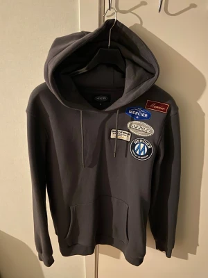 Svart hoodie från Mercier med patches - Svart hoodie från Mercier med flera broderade patches på bröstet i blått, vitt och rött. Klassisk känguruficka framtill och justerbar huva med dragsko. Perfekt för en streetwear-look och riktigt skön passform.