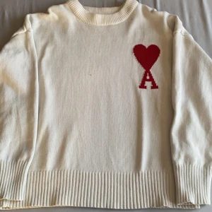 Ami Paris tröja använd bara några gånger  - Säljer en stilren vit stickad tröja med ett rött hjärtmönster och bokstaven 'A' på bröstet. Tröjan har långa ärmar och ribbade kanter vid hals, ärmslut och nederkant. Perfekt för en mysig och trendig look.