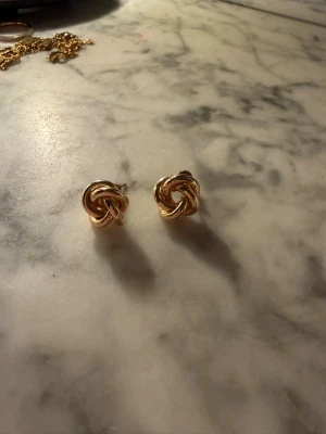 Guldiga knutörhängen i metall - Säljer ett par stilrena örhängen i form av knutar i guldfärgad metall. Perfekta för dig som gillar en klassisk och trendig look. Örhängena är små och diskreta men ändå unika i sin design.