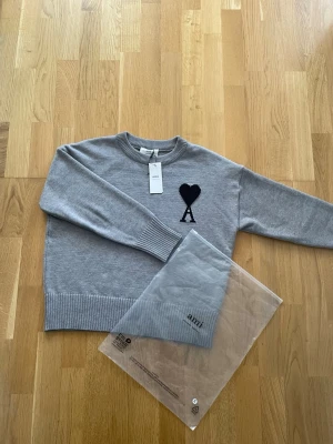 Grå stickad tröja från Ami - Snygg grå stickad tröja från Ami Alexandre Mattiussi med svart hjärta och A-logga på bröstet. Tröjan har rund hals, ribbstickade muddar och nederkant. Perfekt för dig som gillar stilrena och trendiga plagg.