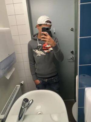 Grå Superdry hoodie med tryck - Snygg grå hoodie från Superdry med stort tryck i vitt, rött och svart på bröstet. Klassisk känguruficka och vit dragsko i huvan. Perfekt för en avslappnad streetwear-look. Passar dig som bäst vid 165 cm lång!