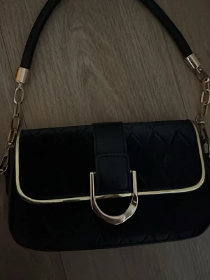 Svart quiltad handväska med gulddetaljer - Stilren svart handväska med quiltat mönster på framsidan, guldfärgade detaljer och kedja. Väskan har en bred axelrem med kedjelänkar och en stor dekorativ guldfärgad spänne framtill. Perfekt accessoar för att lyfta din outfit.