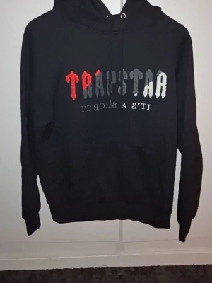 Svart hoodie från Trapstar - Svart hoodie från Trapstar med tryck i rött, grått och vitt på bröstet där det står 'Trapstar It's a secret'. Klassisk huva med dragsko och känguruficka framtill. Perfekt för dig som gillar streetwear och vill sticka ut med en snygg logga.