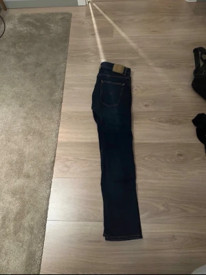 Mörkblå jeans från Nudie Jeans - Säljer ett par mörkblå jeans från Nudie Jeans med klassisk femficksdesign och kontrastsömmar. Jeansen är använda två gånger.