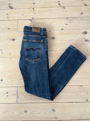 Blå dry denim jeans från Nudie  - Säljer ett par klassiska blå dry denim jeans från Nudie Jeans med raka ben och snygga slitningar. Modell grim Tim storlek 31W 32L men skulle säga att de sitter som en 29/30W. De är inte riktigt som selvedge men de får fortfarande tydliga och fina slitningar. Jeansen har fem fickor, kontrastsömmar och en patch i brunt läder baktill i midjan. Perfekta för en avslappnad och trendig look. Sjukt bra skick och vid frågor är det bara att säga till Mvh Olof.