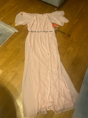 Ljusrosa långklänning med pärldetaljer - Elegant ljusrosa lite beige aldrig långklänning med offshoulder-ärmar och vacker pärl- och stenutsmyckning i midjan. Klänningen har ett flytande fall och är tillverkad i ett tunt, lätt material som ger en romantisk känsla. Perfekt för speciella tillfällen. Och är öppen vid benen.