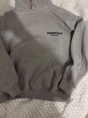 Grå Essentials Fear of God hoodie - Snygg grå hoodie från Fear of God Essentials med svart logga på bröstet. Klassisk passform med huva och stor magficka. Tillverkad i mjukt bomullsmaterial som är perfekt för en avslappnad streetwear-look.