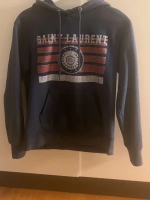 Mörkblå Saint Laurent hoodie - Snygg mörkblå hoodie från Saint Laurent med tryck i vitt och rött framtill. Klassisk känguruficka, justerbar huva med snören och mjukt material. 