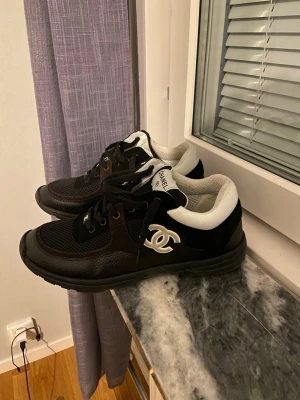 Svarta Chanel sneakers med vit logga - Snygga svarta sneakers från Chanel med vit logga på sidan och vita detaljer runt hälen. Skorna har svart mesh och läder på ovandelen, svarta skosnören och en chunky sula. Perfekta för dig som vill ha en exklusiv och trendig look.