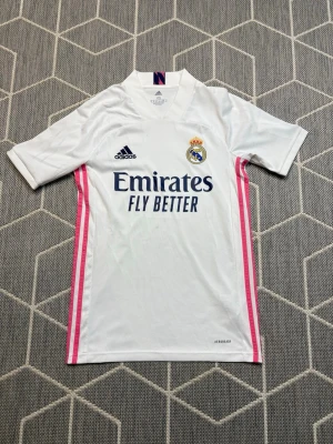 Real Madrid Adidas  - Snygg vit Real Madrid matchtröja från Adidas med klubbmärke och Adidas-logga på bröstet. Tröjan har rosa ränder längs sidorna och sponsortryck 'Emirates Fly Better' på framsidan. Tillverkad i lätt och ventilerande material, perfekt för fotboll eller träning.