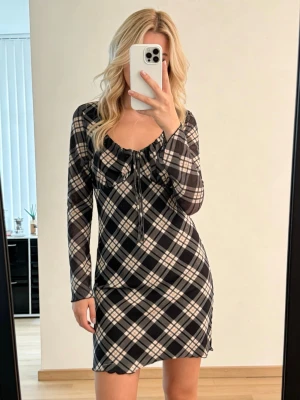 Rutig svartvit kortklänning med snörning - Trendig kortklänning med svartvitt rutmönster och långa ärmar. Klänningen har en snygg snörningsdetalj vid bysten och en figurnära passform. Perfekt för dig som gillar en stilren och modern look.