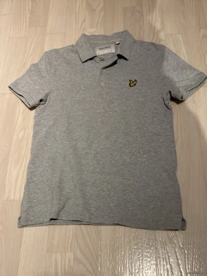 Grå piké från Lyle & Scott - Säljer en stilren grå pikétröja från Lyle & Scott med klassisk krage och två knappar framtill. Tröjan har korta ärmar och den ikoniska gula loggan broderad på bröstet. Tillverkad i mjuk bomull för skön känsla.