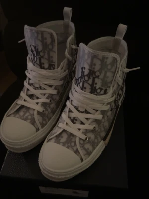 Dior B23 vita high-top sneakers - Säljer ett par Dior B23 high-top sneakers i vitt och grått med ikoniskt Dior-monogram över hela skon. Skorna har genomskinliga paneler, vita snören och robust gummisula. Perfekt för dig som vill sticka ut med exklusiva detaljer och modern design. Pris kan diskuteras 